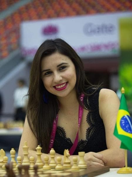 Thauane Medeiros ao tabuleiro na 42ª Olimpíada de Xadrez, com a bandeira do Brasil ao lado